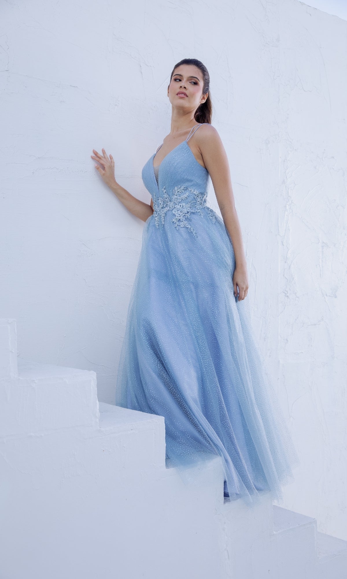 Woman in the Eureka 9797 Lace-Up Long Shimmer Prom Dress stands on white steps against a white textured wall.